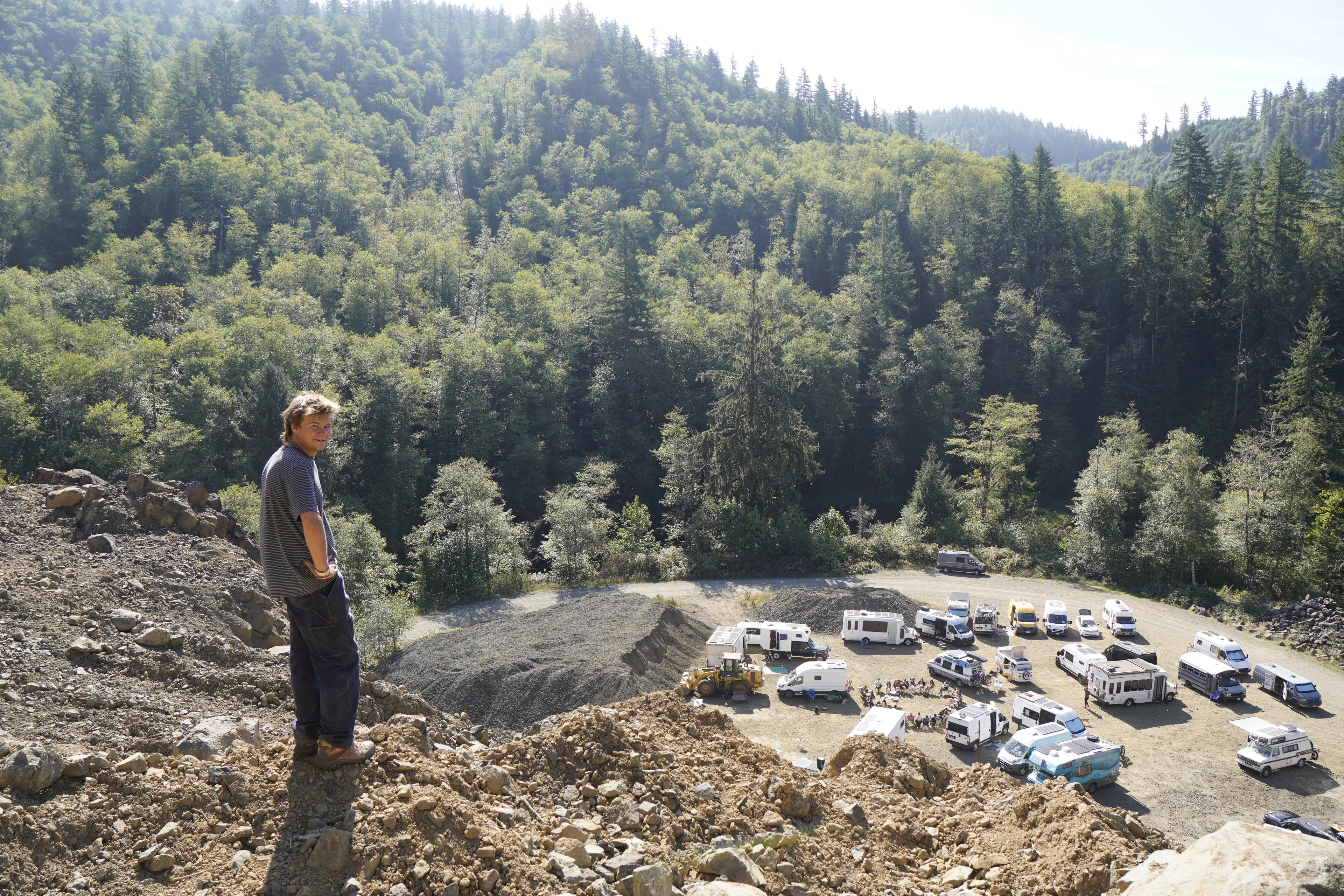 KyleGoesOff standing on a rocky outcrop overlooking a forested area with vehicles parked below