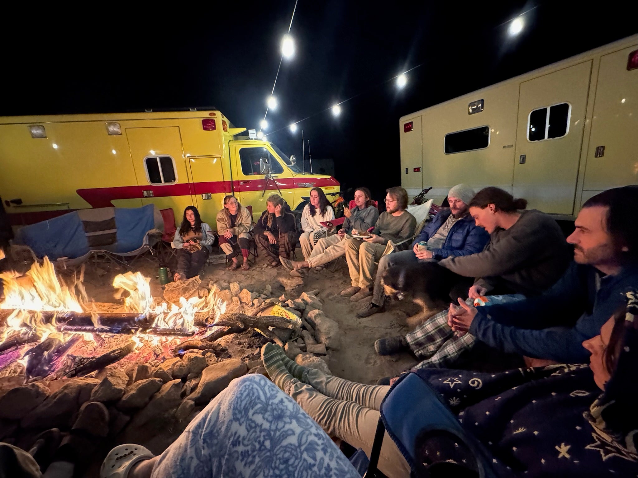 Group of people sitting around a campfire at night with vehicles in the background.