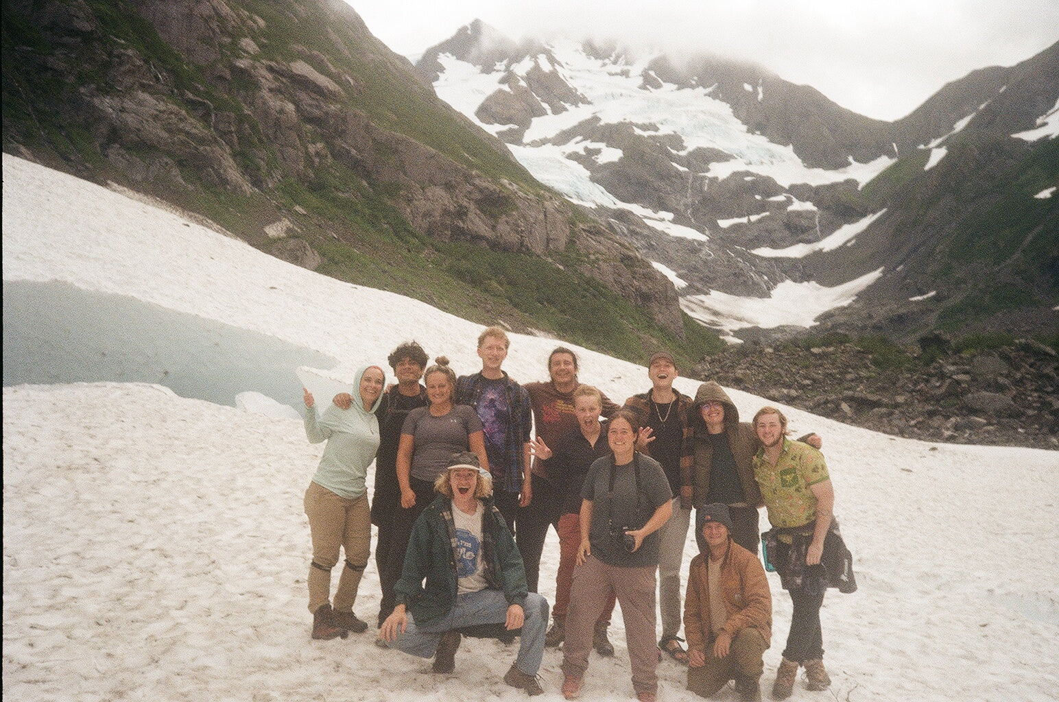Group of people posing for a photo in a mountainous area with snow and greenery.